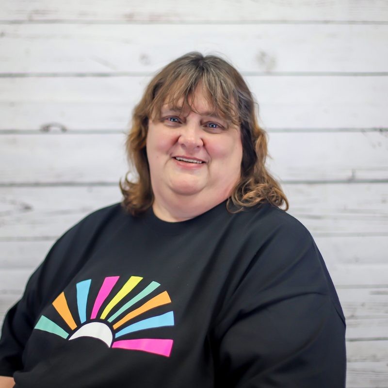 cheryl berg headshot photo with white wood background, bonnie has light brown hair and a CI Therapies black t-shirt on.