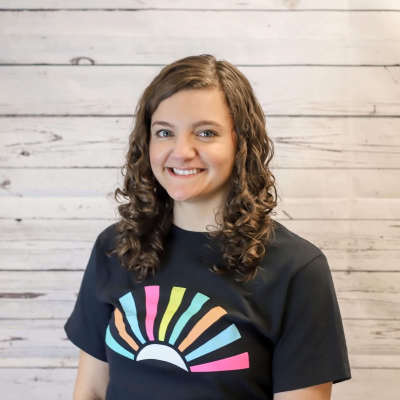 Hannah Henrikson staff biography photo, white wood wall background, hannah has brown curly hair and a black CI Therapies t-shirt on