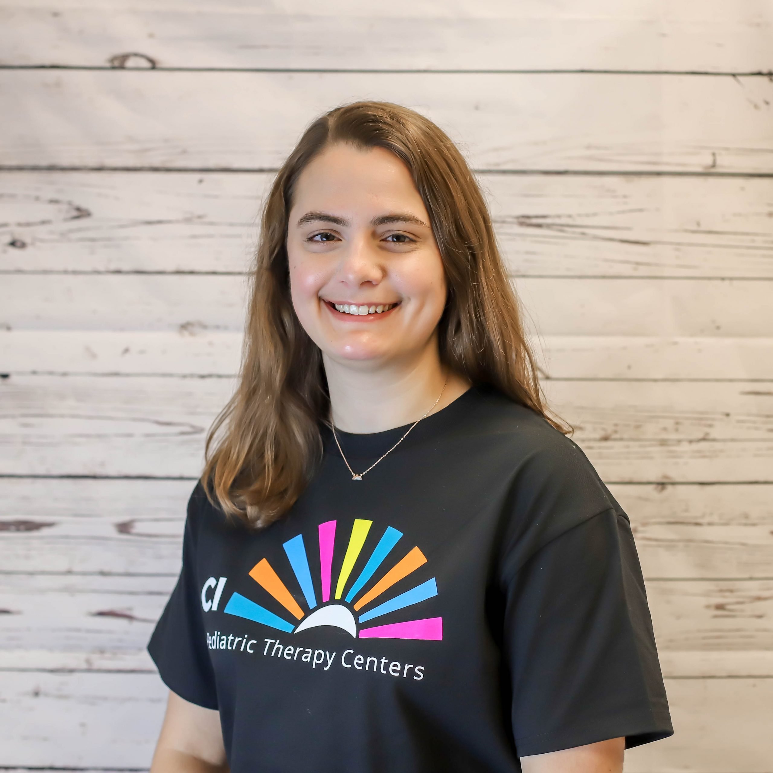 Michelle Hecimovich headshot photo with white wood background, bonnie has light brown hair and a CI Therapies black t-shirt on.
